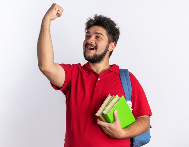 Junger bärtiger Student in rotem Polohemd mit Rucksack, der Notizbücher hält, die glücklich und aufgeregt die Faust ballen und sich über seinen Erfolg freuen, der über weißer Wand steht?