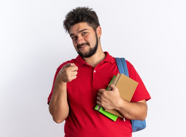 Junger bärtiger Student in rotem Polohemd mit Rucksack, der Notizbücher hält, die mit dem Zeigefinger glücklich und positiv lächelnd über weißer Wand stehen?