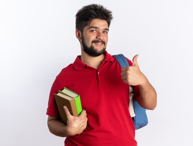 Junger bärtiger Student in rotem Polohemd mit Rucksack, der Notizbücher hält und fröhlich lächelt und Daumen nach oben über weißer Wand zeigt?