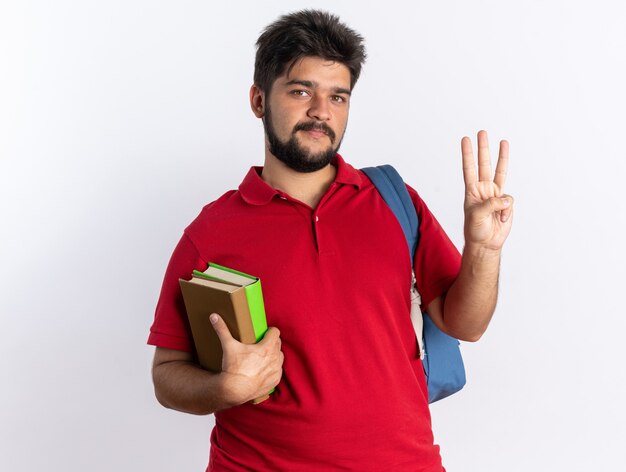 Junger bärtiger Student in rotem Polohemd mit Rucksack, der Notizbücher hält und lächelnd aussieht und Nummer drei mit den Fingern glücklich und positiv steht
