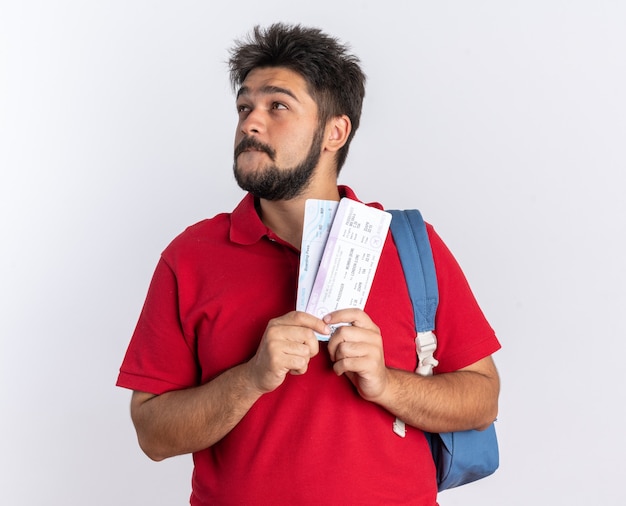 Junger bärtiger Student in rotem Poloshirt mit Rucksack, der Flugtickets hält und mit einem Lächeln im Gesicht aufschaut und positives Ansehen denkt