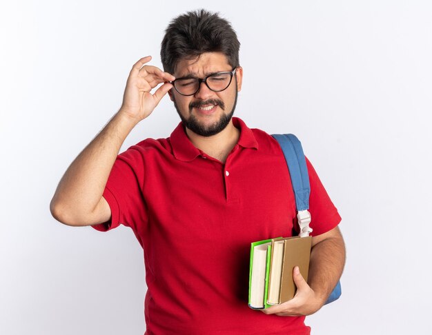 Junger bärtiger Student in rotem Poloshirt mit Rucksack, der Notebooks mit Brille trägt, die glücklich und fröhlich über der weißen Wand stehen?