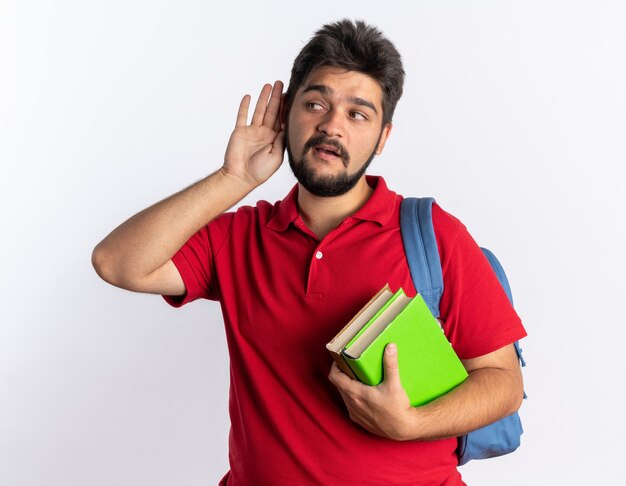 Junger bärtiger Student in rotem Poloshirt mit Rucksack, der Notizbücher hält und die Hand über sein Ohr hält und versucht, zuzuhören und fasziniert zu stehen