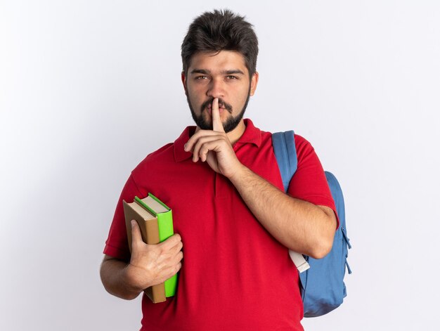 Junger bärtiger Student in rotem Poloshirt mit Rucksack, der Notizbücher hält und die Stille mit dem Finger auf den Lippen stehend macht