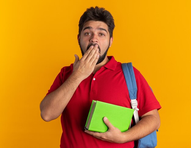 Junger bärtiger Student in rotem Poloshirt mit Rucksack, der Notizbücher hält und schockiert aussieht und den Mund mit der Hand stehend bedeckt