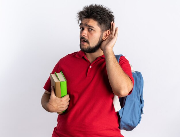 Junger bärtiger Student in rotem Poloshirt mit Rucksack, der Notizbücher hält und versucht, mit der Hand am Ohr zuzuhören