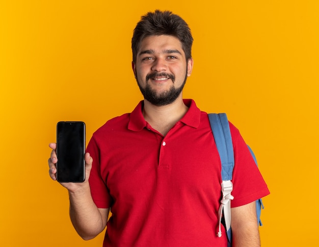 Junger bärtiger Student in rotem Poloshirt mit Rucksack, der Smartphone hält und lächelnd selbstbewusst glücklich und positiv aussieht