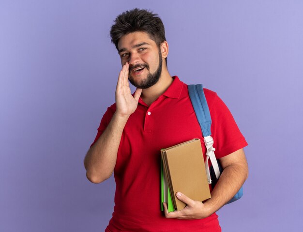 Junger bärtiger Student Kerl im roten Poloshirt mit Rucksack, der Bücher hält, die Kamera betrachten, die ein Geheimnis mit Hand nahe Mund glücklich und positiv steht über blauem Hintergrund flüstert