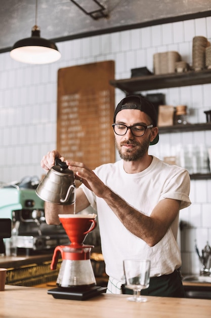 Junger Barista mit Brille und Mütze steht an der Theke und bereitet Kaffee zu, während er im Café arbeitet