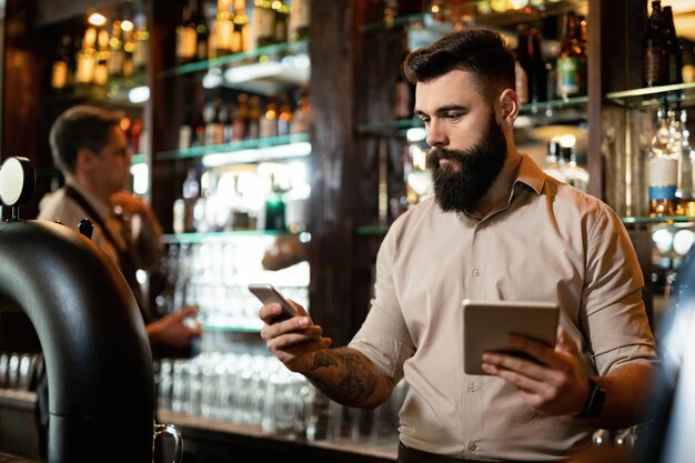 Junger Barkeeper, der Textnachrichten auf dem Handy eingibt, während er ein digitales Tablet in einer Bar verwendet