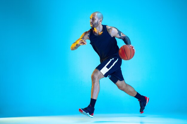 Junger Basketballspieler des Teams, das Sportbekleidungstraining trägt, das in Aktion übt, Bewegung lokalisiert auf blauer Wand im Neonlicht. Konzept von Sport, Bewegung, Energie und dynamischem, gesundem Lebensstil.