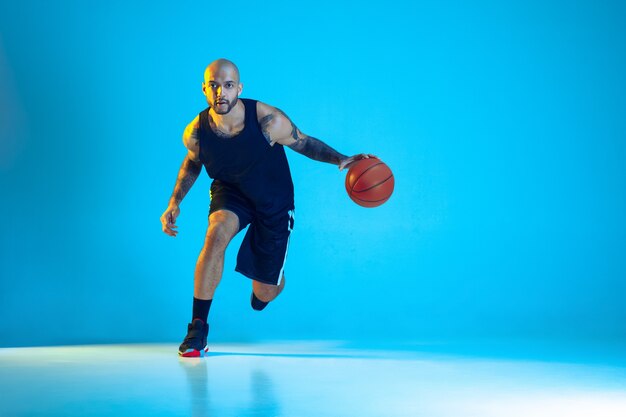 Junger Basketballspieler des Teams, das Sportbekleidungstraining trägt, das in Aktion übt, Bewegung lokalisiert auf blauer Wand im Neonlicht. Konzept von Sport, Bewegung, Energie und dynamischem, gesundem Lebensstil.