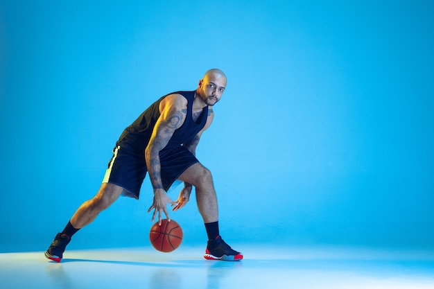 Junger Basketballspieler des Teams, das Sportbekleidungstraining trägt, das in Aktion übt, Bewegung lokalisiert auf blauer Wand im Neonlicht. Konzept von Sport, Bewegung, Energie und dynamischem, gesundem Lebensstil.