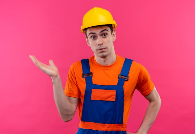 Junger Baumeister, der Bauuniform und Schutzhelm trägt, hebt seine rechte Hand