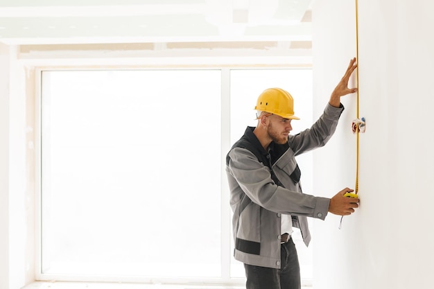 Junger Baumeister in Arbeitskleidung und gelbem Helm mit Maßband bei der Reparatur einer Wohnung mit großem Fenster im Hintergrund