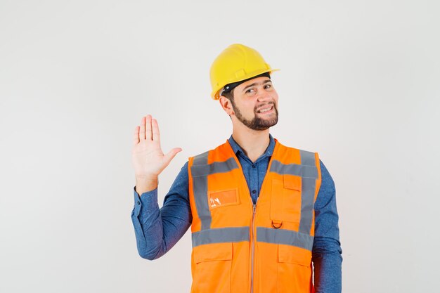 Junger Baumeister winkt mit der Hand, um Hallo oder Auf Wiedersehen in Hemd, Weste, Helm zu sagen und froh auszusehen. Vorderansicht.