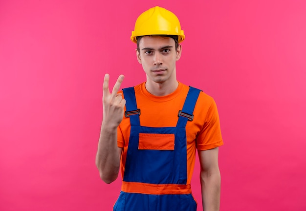 Junger Baumeistermann, der Bauuniform und Schutzhelm trägt, zeigt Friedens- oder Siegesgeste