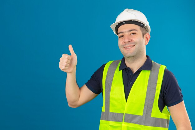 Junger Baumeistermann, der Bauuniform und Sicherheitshelm auf blau lokalisiertem lächelndem positivem tut, das glückliche Daumen hoch Geste mit Hand tut