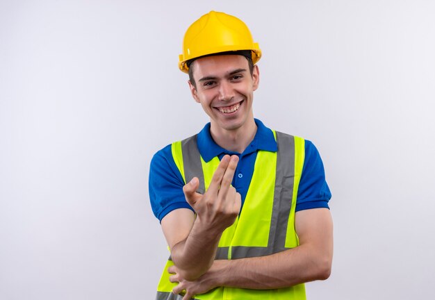 Junger Baumeistermann, der Bauuniform und Sicherheitshelm trägt, lächelt und zeigt Finger
