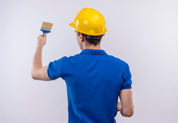 Junger Baumeistermann, der Bauuniform und Sicherheitshelm trägt, malt die Wand mit Pinsel
