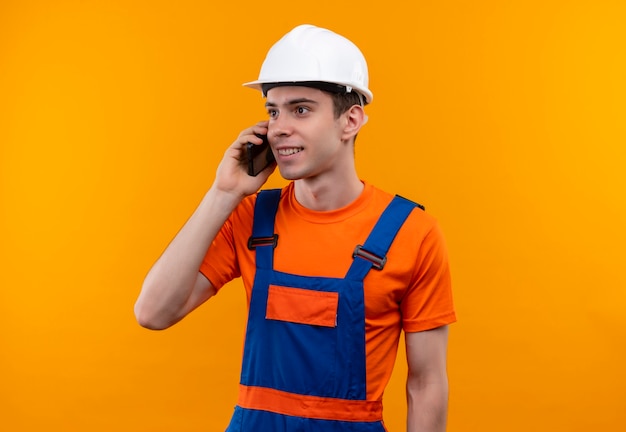 Junger Baumeistermann, der Bauuniform und Sicherheitshelm trägt, spricht glücklich am Telefon