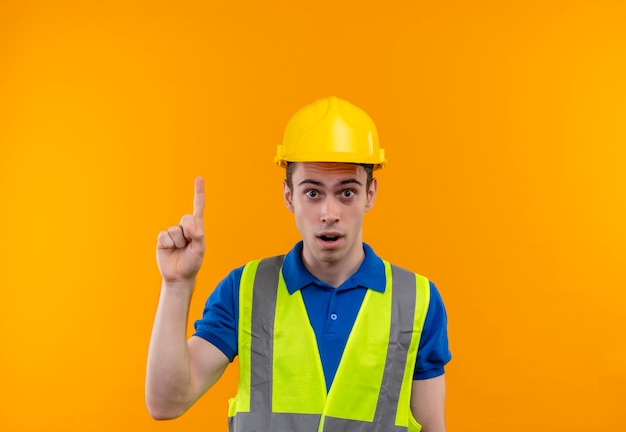 Junger Baumeistermann, der Bauuniform und Sicherheitshelm trägt, zeigt mit überraschtem Finger nach oben