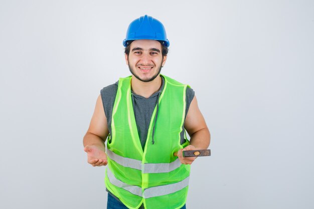 Junger Baumeistermann, der Hammer hält, während Palme in Arbeitskleidunguniform beiseite spreizend und freudig, Vorderansicht schauend.