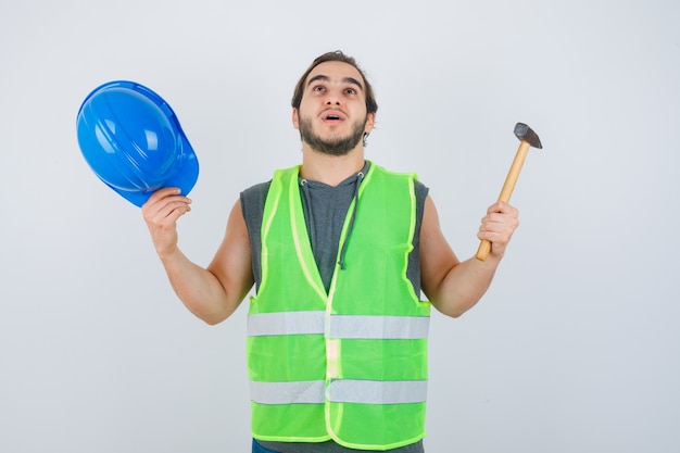 Junger Baumeistermann, der Hammer und Helm hält, während Hände in Arbeitskleidunguniform hebt und ratlos, Vorderansicht schaut.