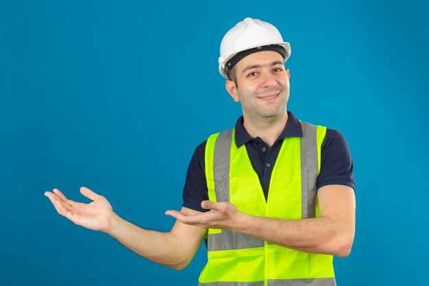 Junger Baumeistermann, der weißen Helm und eine gelbe Weste trägt, mit einem Lächeln auf Gesicht, das mit Handfläche auf Kopienraum auf Blau lokalisiert zeigt