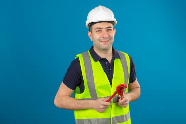 Junger Baumeistermann, der weißen Helm und eine gelbe Weste trägt, Schraubenschlüsselwerkzeug in der Hand mit einem Lächeln auf Gesicht auf blau lokalisiert hält
