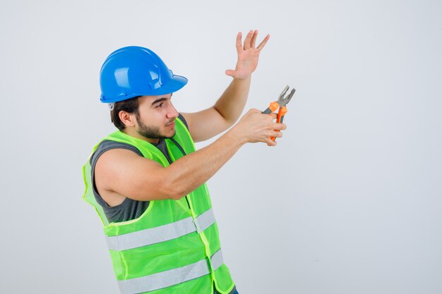 Junger Baumeistermann, der Zangen hält, während Handfläche in Arbeitskleidunguniform angehoben wird und selbstbewusst aussieht. Vorderansicht.