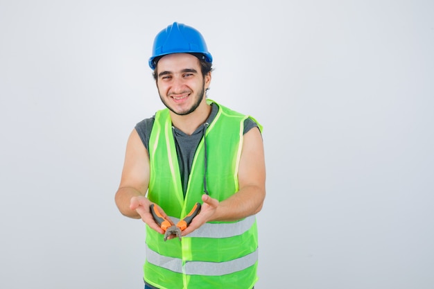 Junger Baumeistermann, der Zangen in Arbeitskleidunguniform zeigt und freudig, Vorderansicht schaut.