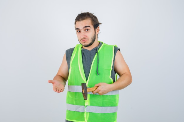 Junger Baumeistermann in Arbeitskleidunguniform, der Hammer hält, während er etwas zeigt und unentschlossen schaut, Vorderansicht.