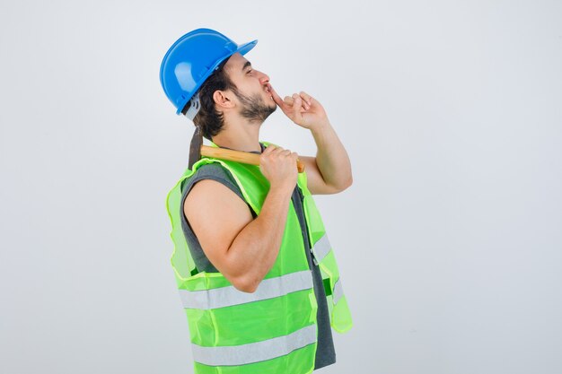 Junger Baumeistermann in Arbeitskleidunguniform, die Hammer auf Schulter hält, während Schweigegeste zeigt und vorsichtig, Vorderansicht schaut.