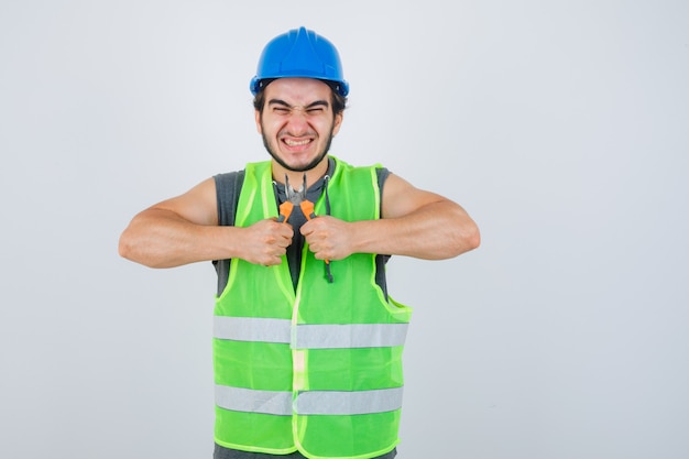 Junger baumeistermann in arbeitskleidunguniform, die zangen hält und glückliche vorderansicht schaut.