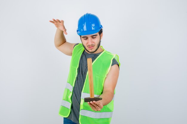 Junger Baumeistermann in der Arbeitskleidunguniform, die Hammer hält, während Hand hebt und vorsichtig schaut, Vorderansicht.