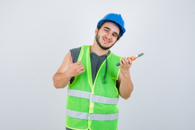 Junger Baumeistermann in der Arbeitskleidunguniform, die Zangen hält, während Daumen oben zeigt und fröhlich, Vorderansicht schaut.