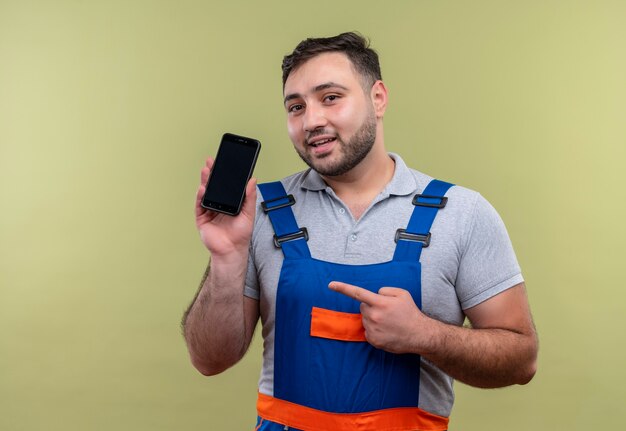 Junger Baumeistermann in der Bauuniform, die das Smartphone zeigt, das mit dem Finger auf es schaut, das sicheres Lächeln über grünem Hintergrund schaut