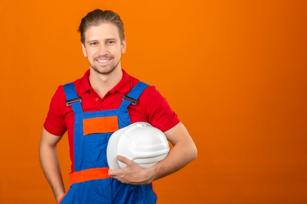 Junger Baumeistermann in der Bauuniform, die Sicherheitshelm in der Hand mit großem Lächeln auf Gesicht hält, das über isolierter orange Wand mit Kopienraum steht