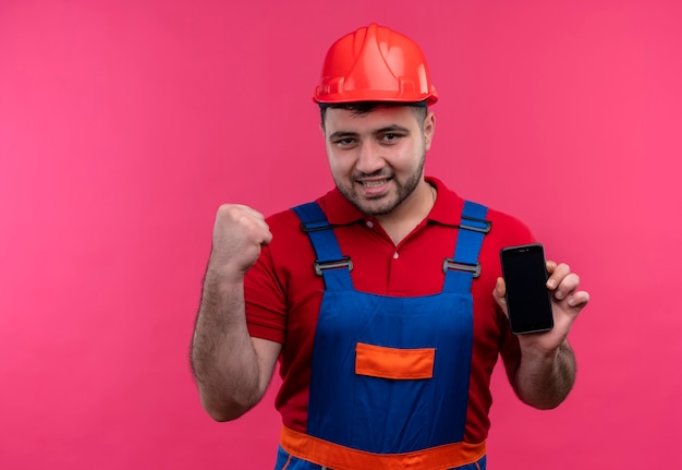 Junger Baumeistermann in der Bauuniform und im Schutzhelm, der die geballte Faust des Smartphones glücklich hält und sich über seinen Erfolg freut