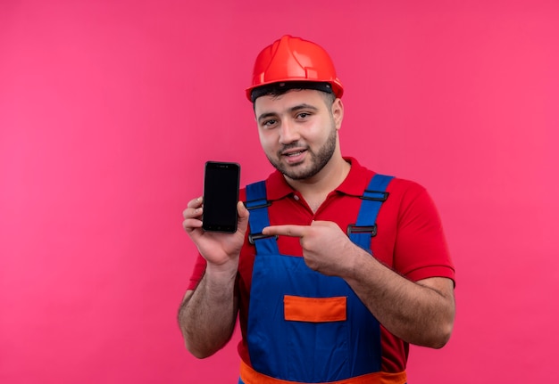 Junger Baumeistermann in der Bauuniform und im Sicherheitshelm, der Smartphone hält, das mit Finger zum int lächeln zeigt