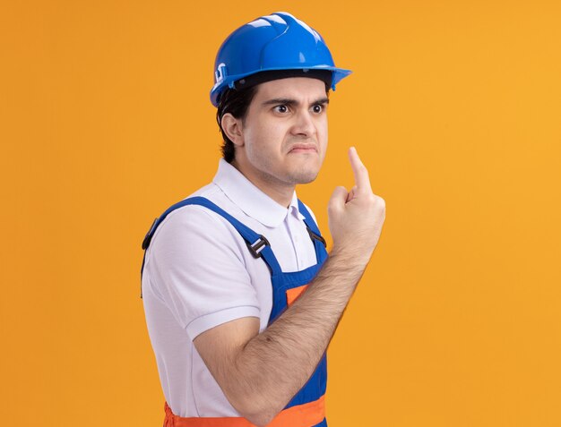 Junger Baumeistermann in der Bauuniform und im Sicherheitshelm, die beiseite schauen mit dem verärgerten Gesicht, das Zeigefingerwarnung über orange Wand steht