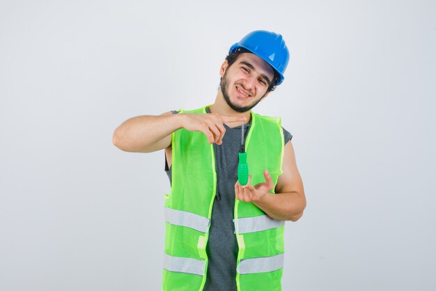 Junger Baumeistermann in der Uniform, die Größenschild mit Schraubendreher zeigt und glückliche Vorderansicht schaut.