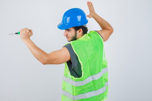 Junger Baumeistermann in der Uniform, die Hand hebt, um mit Schraubendreher zu schlagen und verrückt zu sehen, Vorderansicht.