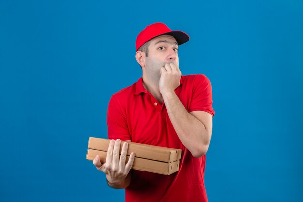 Junger besorgter Lieferbote in der roten Uniform, die Pizzaschachteln hält, die gestresst und nervös mit Händen auf Mund beißen Nägel über lokalisiertem blauem Hintergrund suchen