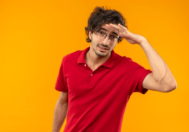 Junger besorgter Mann im roten Hemd mit optischer Brille hält Handfläche an der Stirn, die versucht, etwas entfernt isoliert auf orange Wand zu sehen