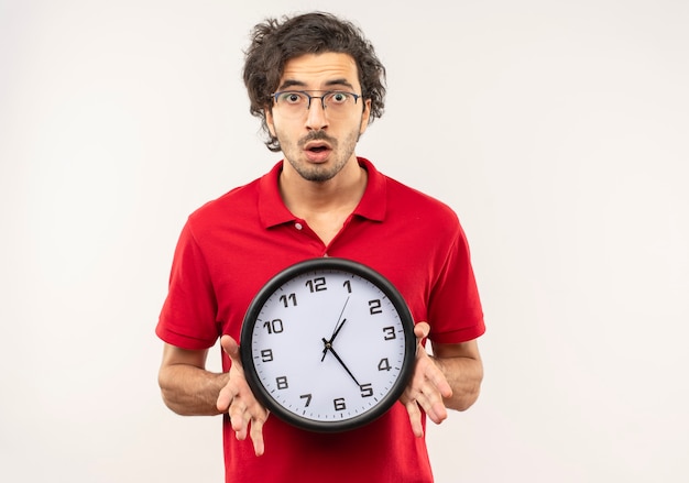 Junger besorgter Mann im roten Hemd mit optischer Brille hält Uhr und schaut lokalisiert auf weißer Wand