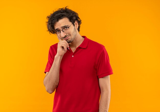 Junger besorgter Mann im roten Hemd mit optischer Brille legt Hand auf Kinn und sieht isoliert auf orange Wand