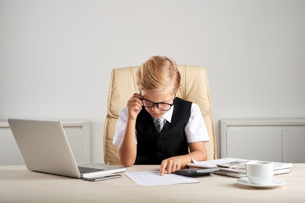 Junger blonder kaukasischer Junge, der am Exekutivschreibtisch im Büro sitzt und Dokumente studiert