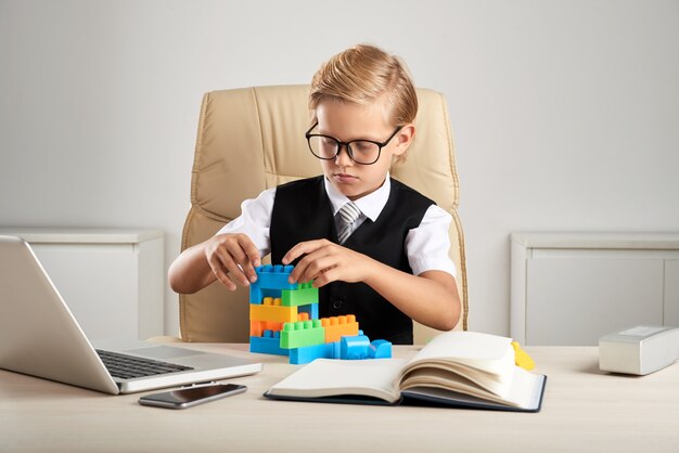 Junger blonder kaukasischer Junge, der im Exekutivstuhl im Büro sitzt und mit Bausteinen spielt